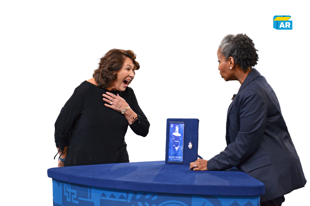 Two women at a blue table on the Antiques Roadshow. The woman on the left is gasping and holding her chest in surprise. The woman on the right, wearing a blazer, is showing her a framed picture and a small item inside a display case.
