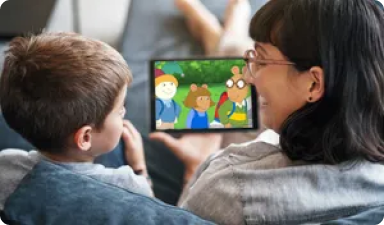 A child and an adult sit on a couch, closely watching a tablet together. The tablet screen displays an animated show featuring anthropomorphic animals. The adult wears glasses and smiles, while the child focuses on the screen, holding it with one hand.