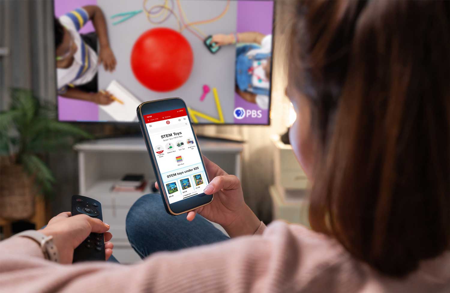 A person is sitting on a couch, holding a smartphone and a TV remote. The smartphone displays a STEM Toys webpage while the TV in the background shows a PBS program featuring children working on a craft project.